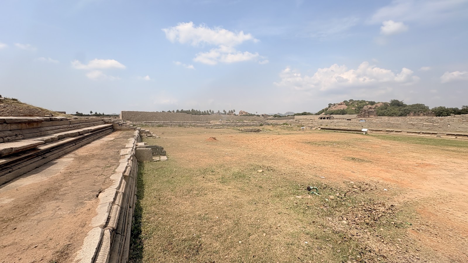 Stone platform base and enclosure walls at Hampi, with open ground and a small shrine on a rocky hill in the distance